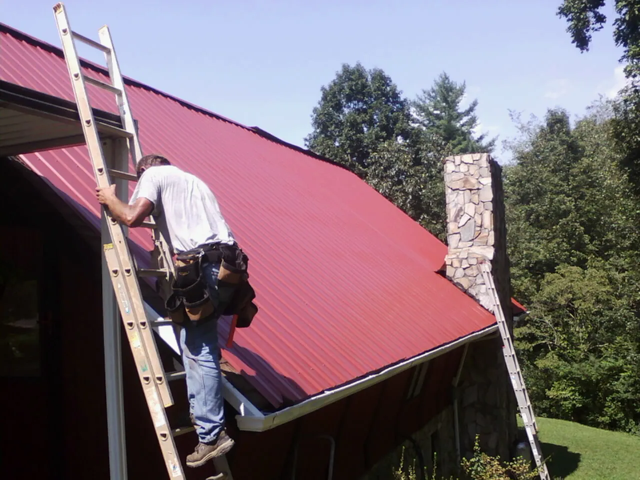 Roofer on a ladder beside a red metal roof and stone chimney — Soffit & Fascia Repair by Roofing Brothers in Cheraw